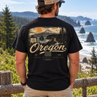 A man wearing a black graphic t-shirt with a vintage-style Oregon landscape print on the back, standing outdoors in front of a wooden fence with a scenic ocean view in the background.