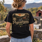 A woman viewed from behind wearing a black graphic t-shirt with a vintage-style Pennsylvania landscape print on the back, set against a scenic outdoor background of a stone bridge and mountains.