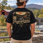 A man viewed from behind wearing a black graphic t-shirt with a vintage-style Pennsylvania landscape print, standing outdoors in front of a stone bridge and mountains.