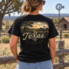 A woman shown from behind wearing a black graphic t-shirt with a vintage-style Texas landscape print on the back, set against a rustic outdoor background with a wooden fence, barn, and windmill.