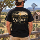 A man viewed from behind wearing a black graphic t-shirt with a large vintage-style Texas landscape print on the back, standing outdoors in front of a rustic fence with a windmill and hills in the background.
