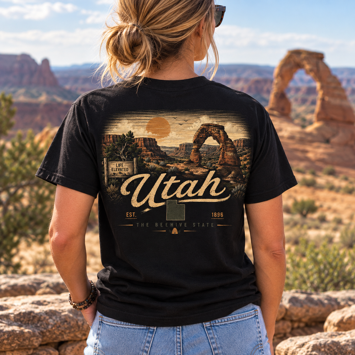 A woman viewed from behind wearing a black graphic t-shirt with a vintage-style Utah desert landscape print featuring Delicate Arch and the text 'Utah The Bountiful State'. The background shows a real outdoor desert canyon environment with Delicate Arch and red rock formations.