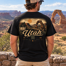 A man viewed from behind wearing a black graphic t-shirt with a large Utah desert landscape print on the back, standing outdoors in front of Delicate Arch National Park.