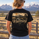 A woman viewed from behind wearing a black graphic t-shirt with a vintage-style Wyoming landscape print, standing outdoors in front of a wooden fence and mountains.