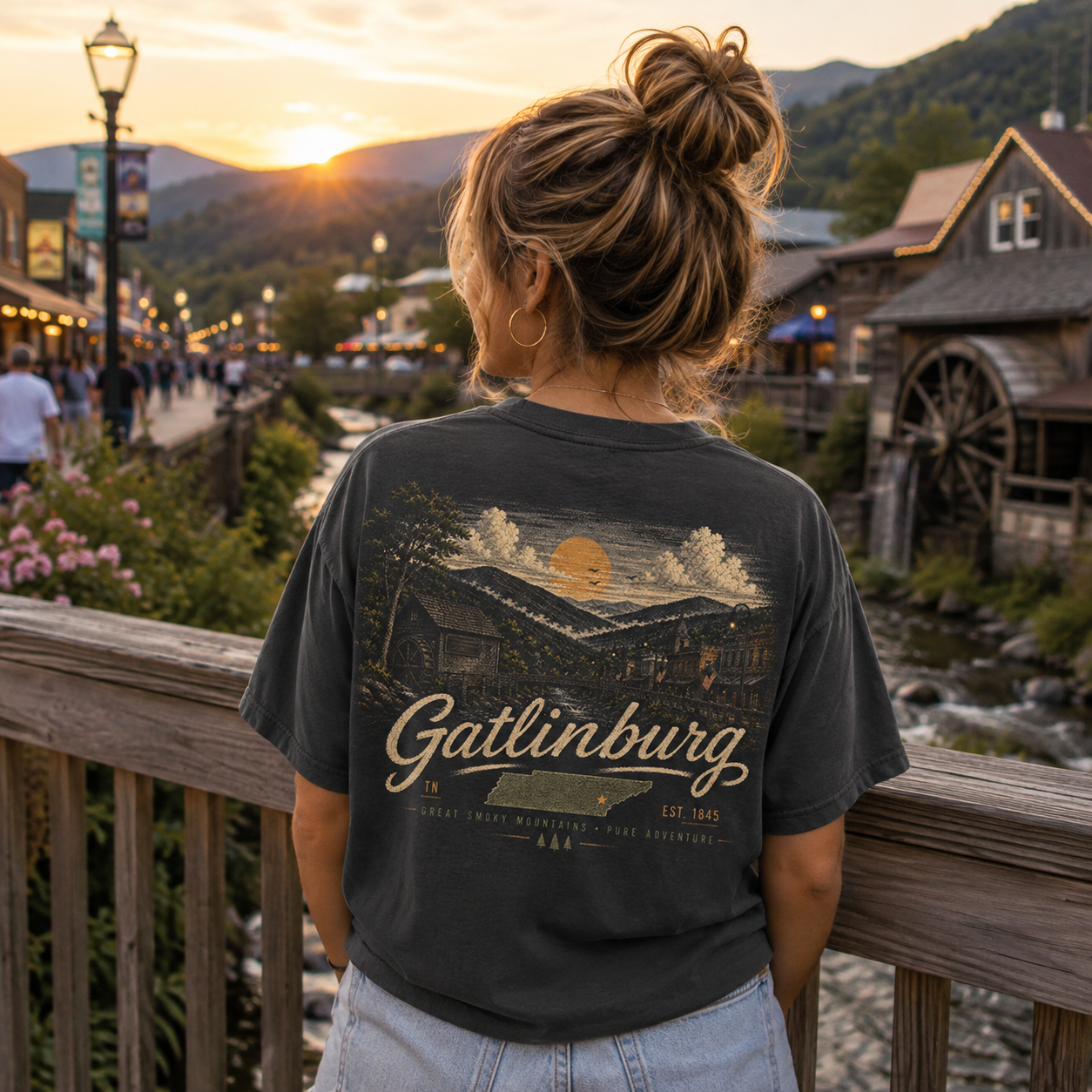 A woman viewed from behind wearing a dark charcoal graphic t-shirt with a scenic Gatlinburg mountain landscape print on the back, standing on a wooden bridge overlooking a mountain town at sunset.