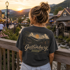 A woman viewed from behind wearing a dark charcoal graphic t-shirt with a scenic Gatlinburg mountain landscape print on the back, standing on a wooden bridge overlooking a mountain town at sunset.