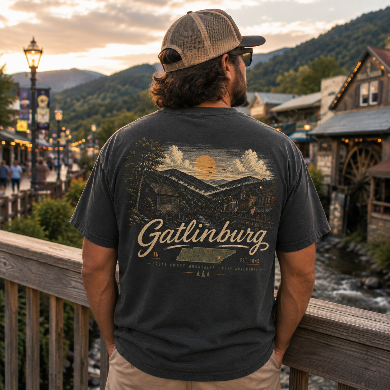 A man wearing a dark charcoal gray t-shirt with a large vintage-style Gattlinburg graphic on the back, standing on a wooden bridge with a scenic mountain town background at sunset.