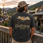 A man wearing a dark charcoal gray t-shirt with a large vintage-style Gattlinburg graphic on the back, standing on a wooden bridge with a scenic mountain town background at sunset.