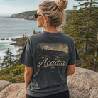 A woman stands outdoors on a rocky overlook with the ocean and pine trees in the background, wearing a dark charcoal graphic T-shirt with a scenic landscape print and the word 'Acadia' across the back.
