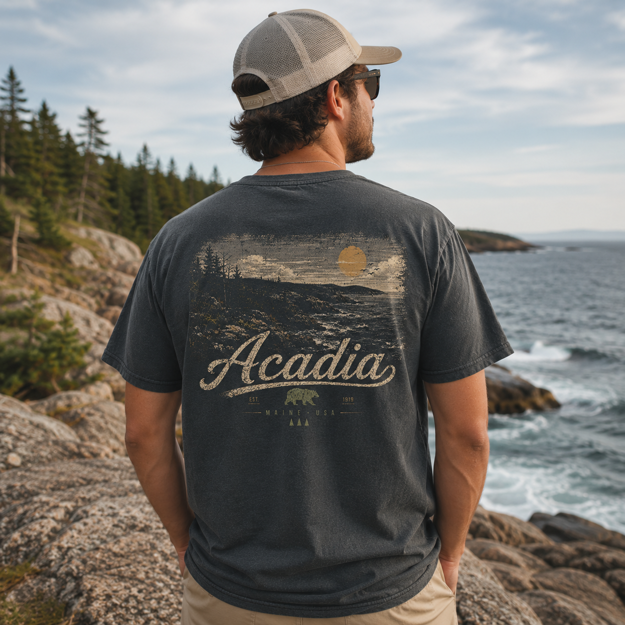 A man wearing a dark charcoal gray t-shirt with a vintage-style coastal graphic that reads 'Acadia Maine' is seen from behind, looking out over a rocky ocean coastline. The t-shirt features a distressed landscape print with a sun and bear.