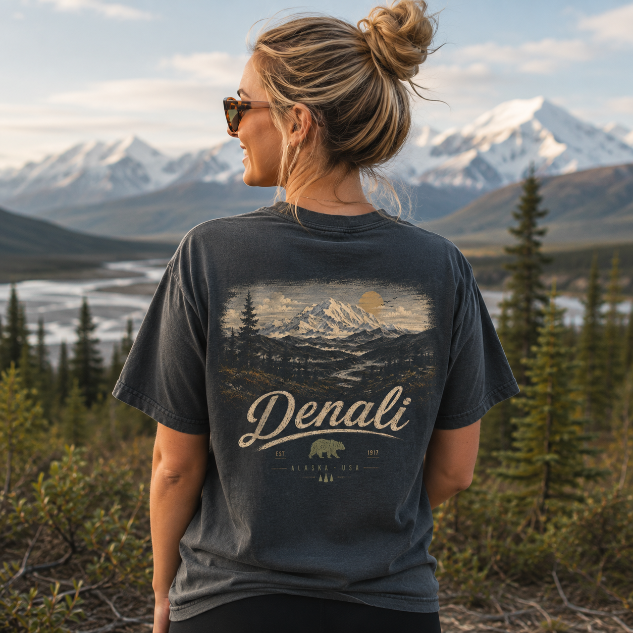 A woman stands outdoors in front of snow-capped mountains and evergreen trees, wearing a dark charcoal graphic T-shirt. The back of the shirt features a scenic mountain print with the word 'Denali' in large script and smaller text reading 'Alaska USA' and '2023'.