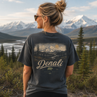 A woman stands outdoors in front of snow-capped mountains and evergreen trees, wearing a dark charcoal graphic T-shirt. The back of the shirt features a scenic mountain print with the word 'Denali' in large script and smaller text reading 'Alaska USA' and '2023'.