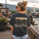 A woman stands on a wooden boardwalk wearing a dark charcoal graphic t-shirt with a mountain landscape print and the words 'Pigeon Forge' on the back. The background shows a scenic outdoor setting with a Ferris wheel, water, and buildings at sunset.