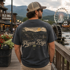 A man wearing a dark charcoal gray t-shirt with a detailed mountain landscape graphic and the text 'Pigeon Forge' on the back, standing on a wooden boardwalk with a rustic town background.