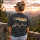 A woman stands on a wooden railing overlooking a mountain landscape at sunset, wearing a dark charcoal gray t-shirt with a large graphic print on the back that reads 'The Great Smokey Mountains' with a bear and mountain scene. She is wearing denim shorts.