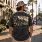 A man wearing a charcoal gray graphic t-shirt with a vintage-style Charleston, SC illustration on the back, standing on a sunny city sidewalk.