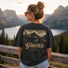 A woman stands outdoors in front of a mountain lake landscape at sunset, wearing a dark charcoal graphic T-shirt with a scenic mountain print and the word "Glacier" on the back, paired with light blue jeans.