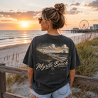 A woman stands on a beach boardwalk at sunset wearing a dark charcoal graphic t-shirt with 'Myrtle Beach SC' printed on the back, paired with light denim shorts.