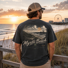 A man stands on a beach boardwalk at sunset wearing a dark charcoal t-shirt with a large back graphic that reads 'Myrtle Beach' and shows the ocean, dunes, and a Ferris wheel in the background.