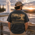 A man stands with his back to the camera wearing a dark charcoal t-shirt with a vintage Yellowstone National Park graphic on the back, set against a scenic sunset landscape with a geyser erupting in the background.