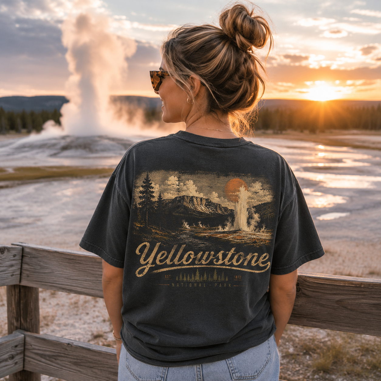 A woman stands outdoors at Yellowstone National Park at sunset, viewed from behind, wearing a dark charcoal graphic T-shirt with a large Yellowstone landscape print and light blue jeans.
