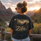 A woman stands outdoors in a mountain landscape at sunset wearing a dark charcoal graphic T-shirt with a large scenic print and the word 'Zion' across the back, paired with light blue denim shorts.
