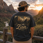 A man stands outdoors in a mountain landscape at sunset, viewed from behind, wearing a dark charcoal T-shirt with a large scenic mountain graphic and the word "Zion" across the back, along with a tan trucker cap and khaki pants.