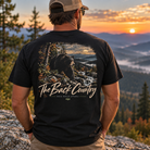 A man wearing a black graphic t-shirt with a bear and mountain landscape print on the back, standing on a rocky mountain peak at sunset. The t-shirt features the text 'The Back Country' and 'BACK DOOR APPAREL'.