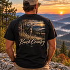 A man viewed from behind wearing a black graphic t-shirt with a large 'BuckCountry' deer hunting scene on the back, set against a scenic mountain sunset background.