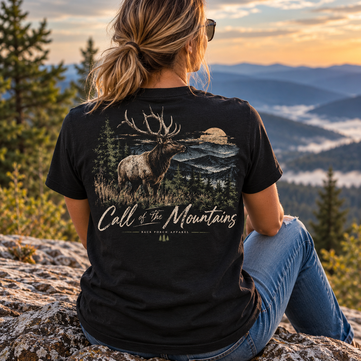 A woman sits on a rock outdoors, viewed from behind, wearing a black graphic T-shirt with a large elk and mountain scene on the back and the words 'Call of the Mountains' and 'Black Forest Apparel.'.