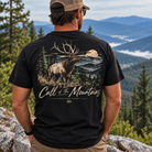 A man viewed from behind wearing a black graphic t-shirt with a large back print of an elk in a mountain landscape and the text 'Call of the Mountains'. He is outdoors on a rocky mountain peak with pine trees and blue mountains in the background.