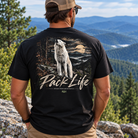 A man wearing a black graphic t-shirt with a large back print of a howling wolf and the text 'Pack Life', photographed from behind against a mountain landscape.