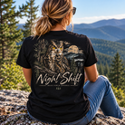 A woman wearing a black graphic t-shirt with a large owl and 'Night Shift' print, sitting outdoors on a mountain peak with a scenic forest and mountain background.