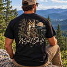 A man wearing a black graphic t-shirt with a large owl and 'Night Shift' print, sitting outdoors on a mountain peak with a scenic forest and mountain background.
