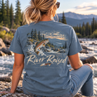 A woman sits on a rock by a river with mountains and trees in the background, wearing a blue-gray t-shirt from the back. The shirt features a large graphic of a jumping fish over water and trees, with the text 'River Raised' and 'Rack Fish Apparel' printed below.
