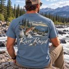 A man sitting outdoors by a rocky river with mountains and pine trees in the background, wearing a blue graphic T-shirt from the back. The shirt features a large fish jumping from the water with the text 'River Raised' and 'Backcountry Apparel' below it.