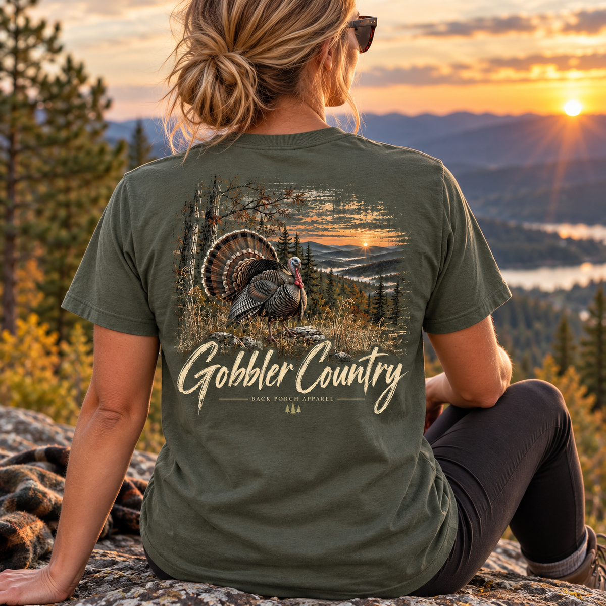 A woman sits on a rock outdoors at sunset wearing an olive green t-shirt with a large back graphic of a turkey in a field and the text 'Gobbler Country' and 'Back Farm Apparel'.