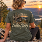 A woman sits on a rock outdoors at sunset wearing an olive green t-shirt with a large back graphic of a turkey in a field and the text 'Gobbler Country' and 'Back Farm Apparel'.