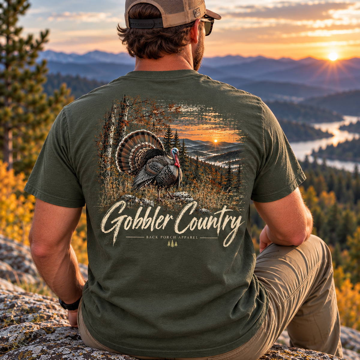A man wearing an olive green t-shirt with a large back graphic of a turkey in a sunset landscape and the text 'Gobbler Country', sitting on a rock overlooking a mountain valley at sunset.
