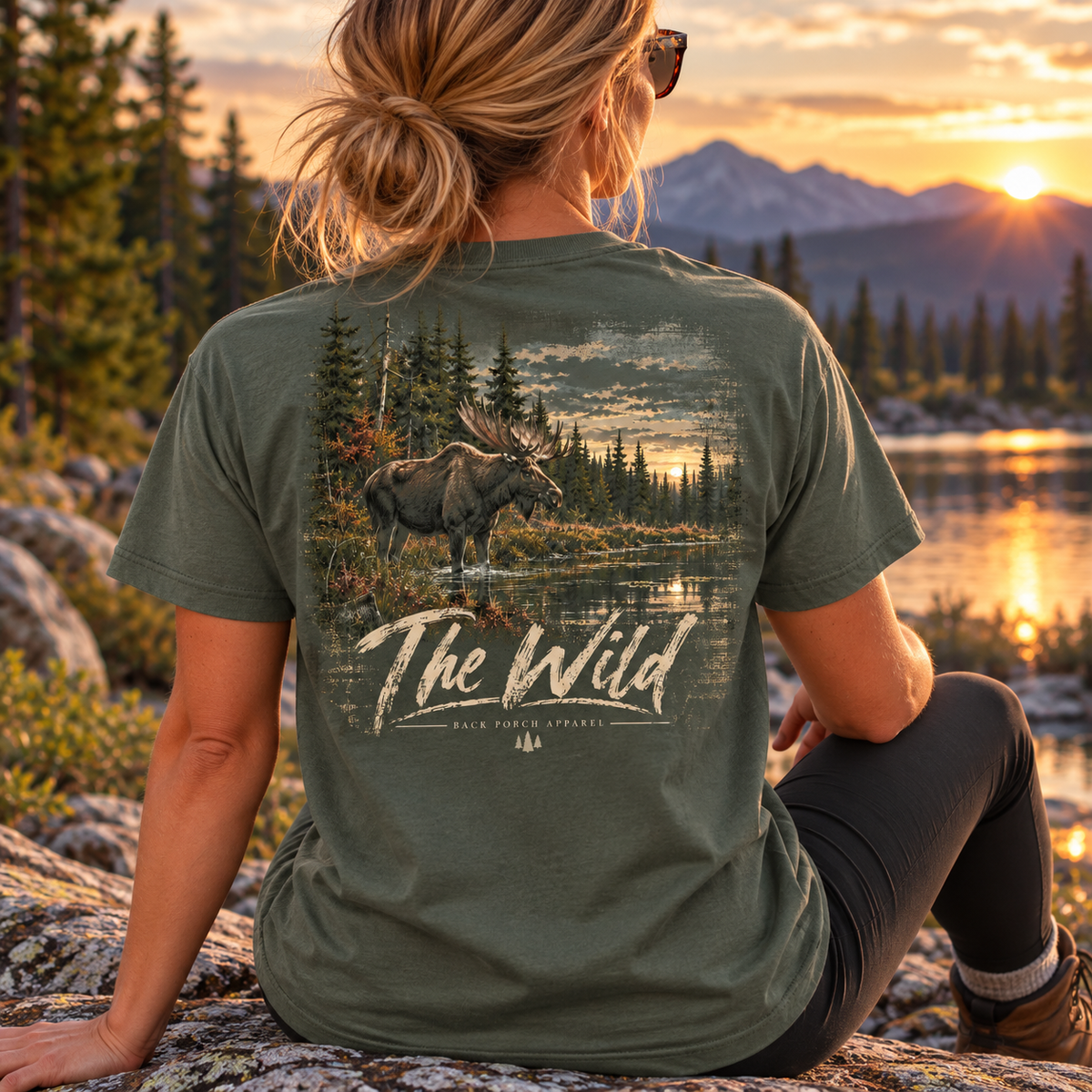 A woman sits outdoors on a rock at sunset facing a mountain lake landscape, wearing an olive green graphic T-shirt with a large back print of a moose in a forest and the words “The Wild.”.