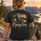 A woman sits on a rocky mountain overlook at sunset wearing a black graphic t-shirt. The back of the shirt features a large graphic of a bald eagle flying over a lake and forest, with the text 'Flying Free' and 'Bald Eagle National Park' printed below.