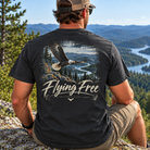 A man sitting outdoors on a rock overlooking a mountain lake landscape, shown from the back wearing a dark charcoal t-shirt with a large graphic of a flying bald eagle and the text 'Flying Free'.