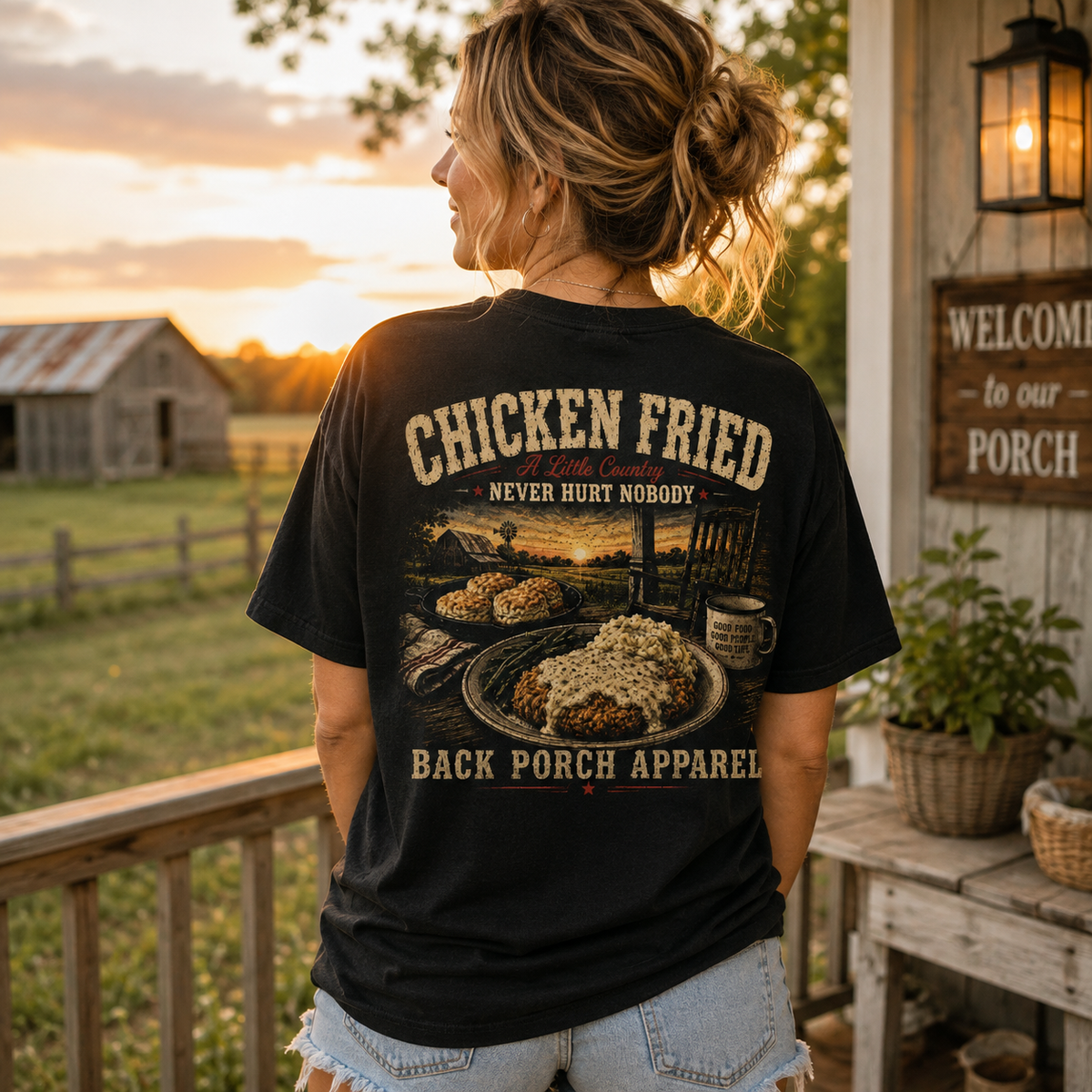 A woman stands on a porch at sunset wearing a black graphic T-shirt. The back of the shirt features a large printed design with the words “CHICKEN FRIED,” “NEVER HURT NOBODY,” and “BACK PORCH APPAREL,” along with an illustration of a plated fried dish and rustic porch imagery.