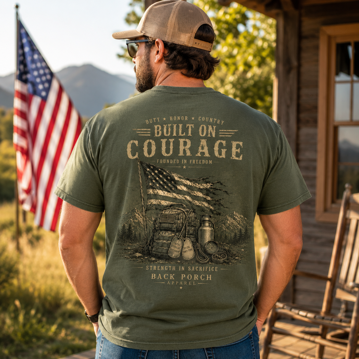 A man wearing an olive green t-shirt with a large vintage-style patriotic graphic on the back, standing outdoors in front of a rustic cabin with an American flag in the background.