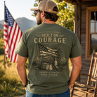 A man wearing an olive green t-shirt with a large vintage-style patriotic graphic on the back, standing outdoors in front of a rustic cabin with an American flag in the background.