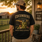 A man stands on a rustic porch at sunset wearing a black graphic T-shirt with a large back print reading “Cucumbers & Onions in Vinegar” and “Back Porch Apparel,” along with a tan cap and khaki pants.