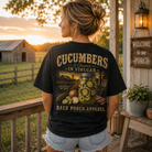 A woman stands on a rustic porch at sunset wearing a black graphic T-shirt with a large back print reading “CUCUMBERS Onions IN VINEGAR” and “BACK PORCH APPAREL,” paired with light denim shorts.