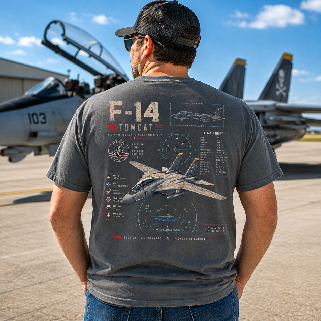 A man viewed from behind wearing a dark gray graphic t-shirt featuring an F-14 Tomcat fighter jet design, standing on an airfield with fighter jets in the background.
