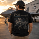 A man viewed from behind wearing a black t-shirt with a detailed F-35 Lightning II graphic print on the back, standing on an airfield tarmac with fighter jets and a hangar in the background at sunset.