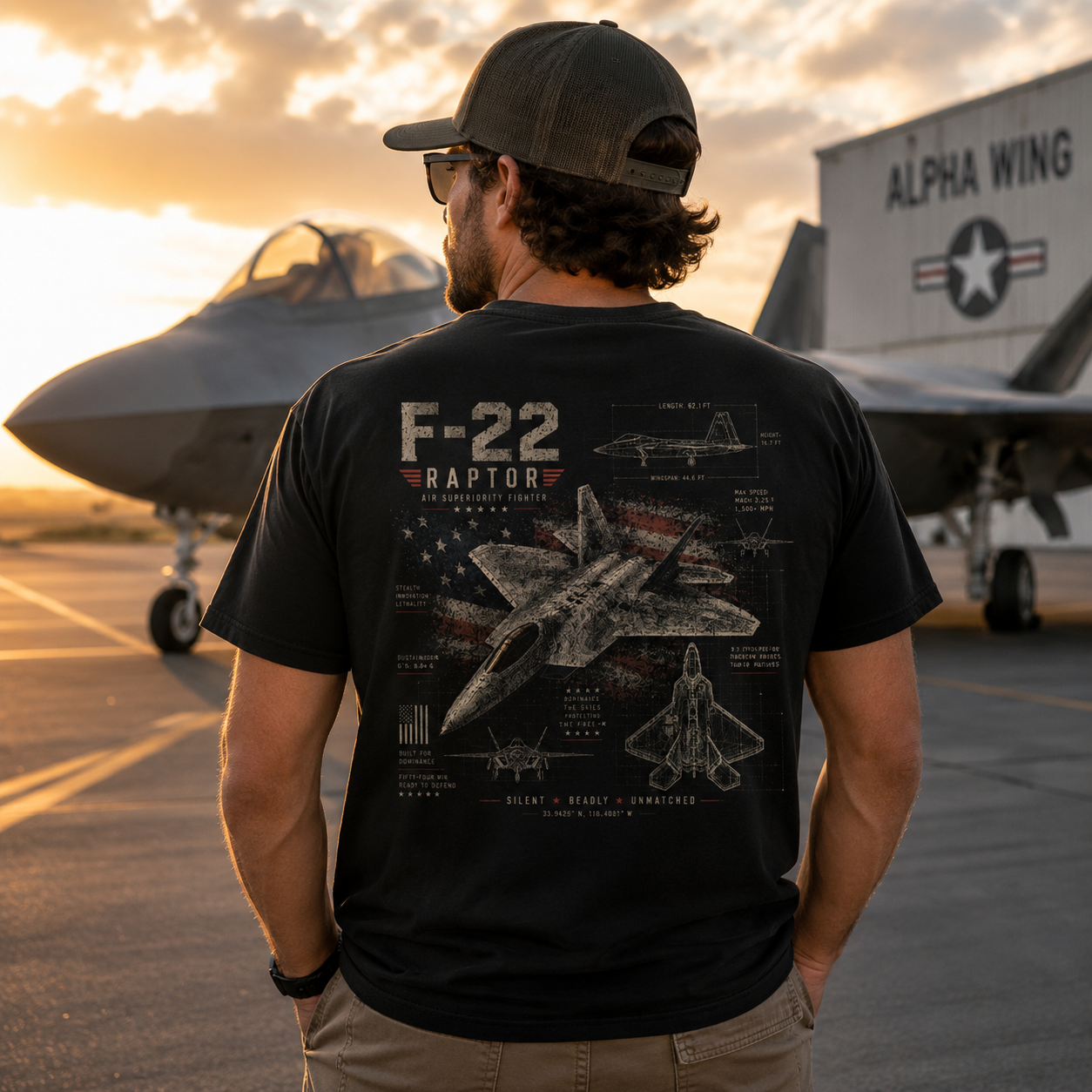 A man viewed from behind wearing a black graphic t-shirt with an F-22 Raptor fighter jet design, standing on an airfield tarmac with fighter jets and a hangar in the background at sunset.